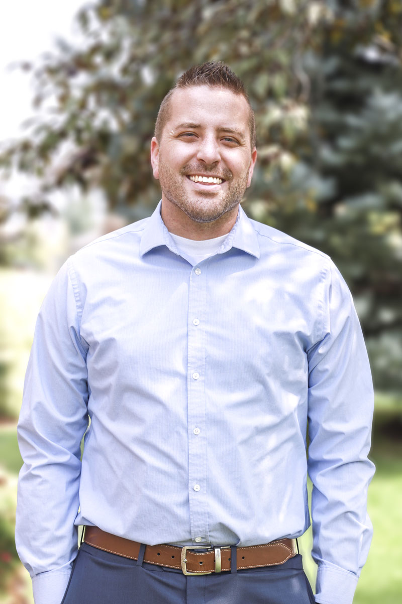 portrait of Chase Hanson, mulat man with dark blonde hair, smiling and wearing light blue shirt, standing outside